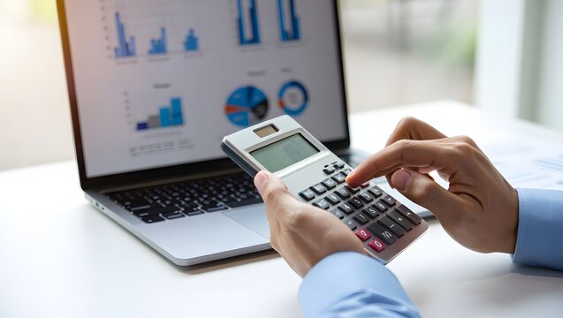 Close up of a person using a calculator while working on a laptop with financial charts