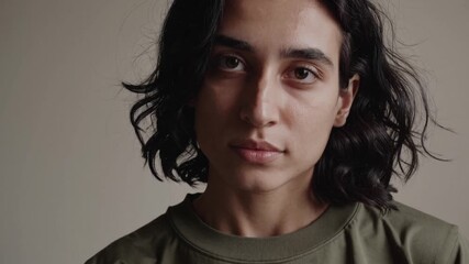 Young hispanic woman with wavy black hair wearing green t shirt standing in calm, contemplative pose against neutral background, gazing directly at camera with serene, introspective expression - Powered by Adobe