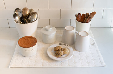 A kitchen still life featuring white coffee cups, utensils and chocolate chip cookies.