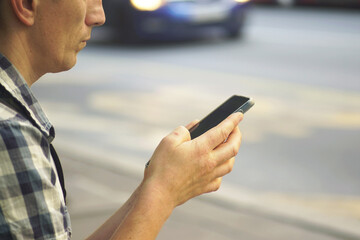Man standing by the roadside with a phone in hand, close-up. Concept of smart commuting, app-based travel planning, digital maps and city navigation. Capturing the role of technology in transportation