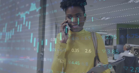 Walking woman in sweater talking on phone and holding folder in office hallway with stock charts
