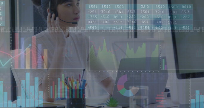Working agent wearing headset with microphone reviewing financial charts at office desk with laptop