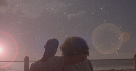 Hugging silhouetted couple gazing over ocean from metal railing under starry sky, with lens flare