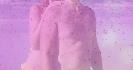 Fototapeta premium Embracing pair wearing dark swim trunks and yellow bikini bottom at beach shoreline, pink overlay