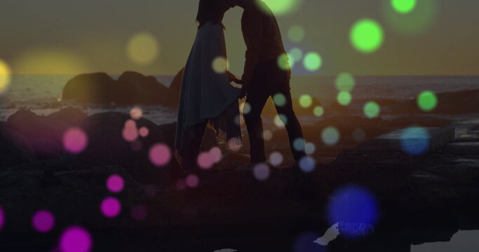 Leaning couple kissing on rocky outcrop with waves at sunset horizon, with decorative bokeh spots