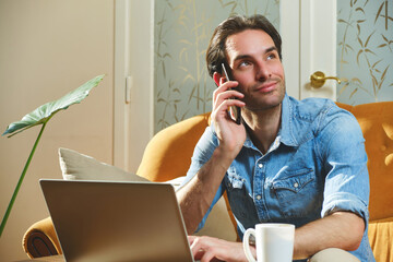 A man is sitting comfortably on a sofa, speaking on his phone with a relaxed expression. A laptop...