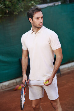 A young man stands confidently on a clay tennis court, holding a racket in one hand and a tennis ball in the other, ready for his next move