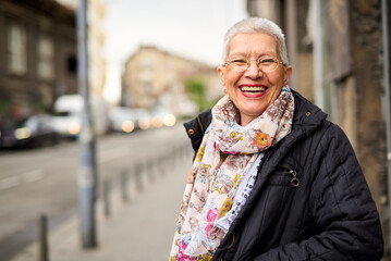 An elderly woman stands on a bustling city street, smiling widely while wearing a stylish scarf....