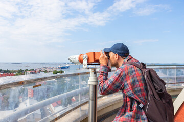 A young male tourist looks through a viewing binocular on an observation deck of Hanko Water Tower, enjoying panoramic views of the cityscape, harbor and coastline on a bright sunny day