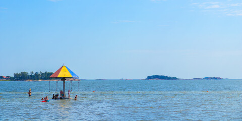 A colorful carousel by the sea on a Hanko beach, Finland, with adults and children enjoying rides...