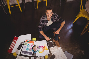 Man sitting cross legged on floor in design studio using smartphone amid design prints and juice