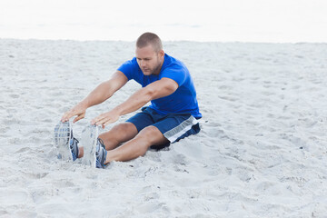 Fit man stretching forward while reaching toward sneakers on sandy beach near shoreline