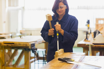 Asian woman wearing goggles and coverall chiseling wood block with mallet, chisel at woodshop bench