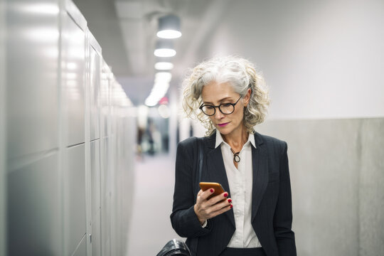 Middle-aged Caucasian businesswoman checking her smartphone while walking past cubicles, wearing elegant suit and glasses, navigating a busy office hallway with efficiency and style