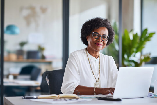 Middle-aged Black business woman participating in a video call with international clients, sitting at her desk with a laptop, dressed in a professional blouse and glasses, her office contemporary with - Powered by Adobe