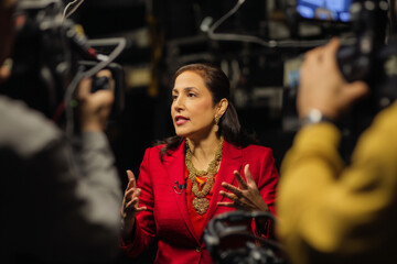 Middle-aged Hispanic businesswoman being interviewed on camera for a business news segment. She wears a red blazer and gold necklace, confidently speaking with gestures while camera crew records.