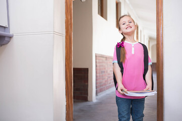 Fototapeta premium Female child standing in wooden door frame in school corridor holding notebooks and backpack