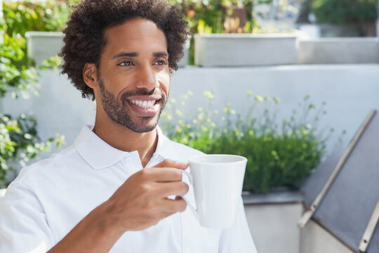African American man wearing white polo shirt and sipping from ceramic mug on terrace with planters