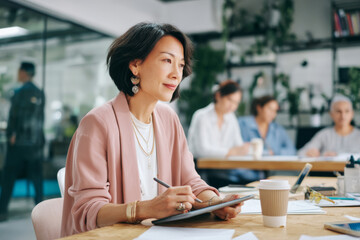 Middle-aged Asian businesswoman sitting in a creative coworking space, writing on a tablet while discussing ideas with colleagues. She's dressed in a pastel blazer, wearing subtle makeup and modern