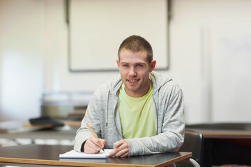 Male student sitting in college classroom writing in notebook with pencil and wood-grain desk