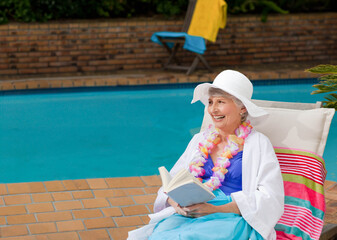 Senior woman wearing sun hat and floral lei reading book on pool patio chair, copy space
