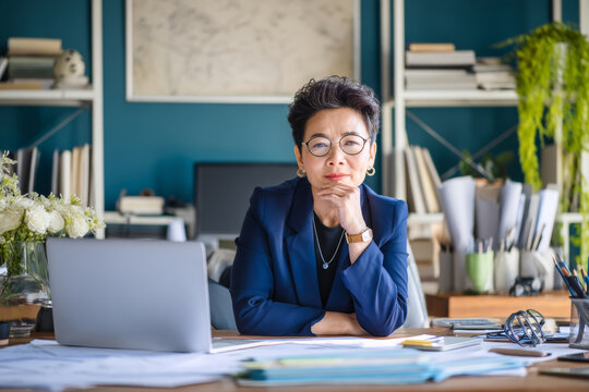 Middle-aged Asian business woman in her office, sitting at a desk covered with project documents and a laptop, engaged in a virtual meeting, wearing a