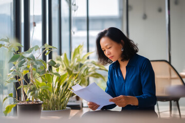 Middle-aged Asian business woman reviewing her team progress reports in her office, dressed in a formal blue blouse, the office well-lit with natural