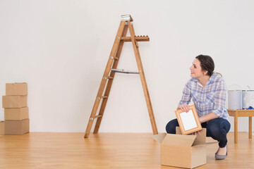 Woman in mid-20s crouching near ladder unpacking picture frame at home with paint cans, copy space