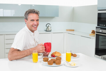 Middle-aged man smiling while holding red mug at home kitchen table with cereal and orange juice