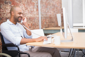 Man in wheelchair holding telephone while typing on keyboard in office, copy space