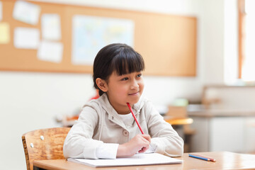 Asian child girl sitting at classroom desk with open notebook holding red pencil near chin
