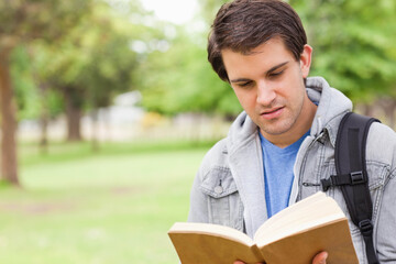 Man standing outdoors in park holding and reading hardcover book with backpack strap, copy space