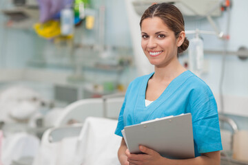 Female nurse holding clipboard while standing in hospital ward with IV stand and beds, copy space
