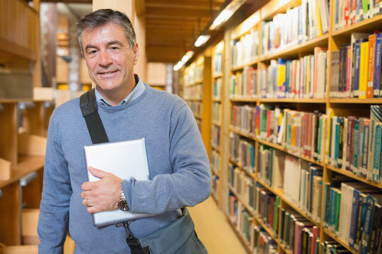 Male in his fifties standing in library aisle holding tablet computer with shoulder bag, copy space