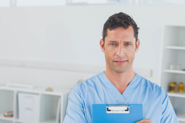 Mature adult man standing in clinical examination room holding blue clipboard with medical supplies