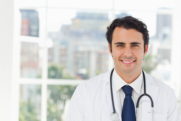 Mid adult male physician standing by office window wearing white coat and stethoscope, copy space