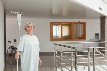 Senior woman holding IV pole in hospital corridor next to wheelchair, wearing gown