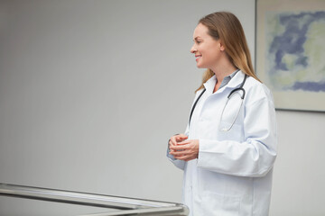 Woman doctor standing beside railing in clinic corridor in lab coat holding stethoscope, copy space