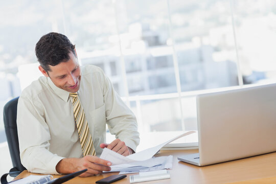 Male office worker in suit flipping through paperwork while sitting at desk in office with laptop