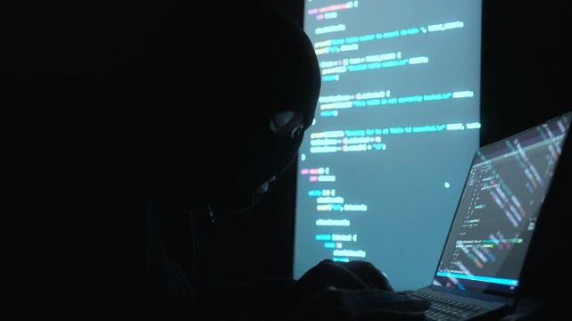 Thief Hacker in balaclava with an obscured face is stealing data, representing cybercrime, hacking, dark web activity, and online security threats in dark room. Cyber Security concept.

