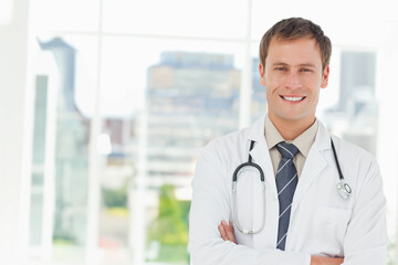 Mid adult male doctor standing arms crossed wearing lab coat and stethoscope in clinic, copy space