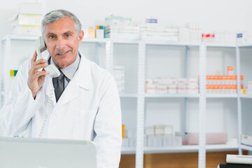 Senior male pharmacist using telephone handset at pharmacy counter with stocked shelves, copy space