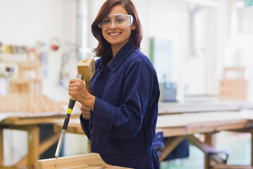 Asian woman wearing navy coveralls and goggles carving block on shop bench with mallet and chisel