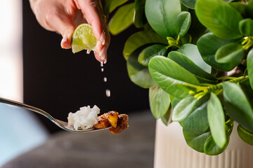 Close-up of a hand squeezing lime juice over a fork with rice and meat, with a green plant in the...