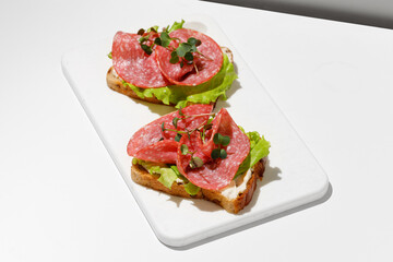 Sandwich, toast with sliced salami, sausage, green lettuce on white cutting board. Hard light,...