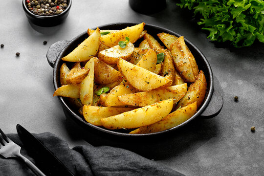 Roasted potatoes with herbs and spices. Baked potato wedges in frying pan on dark stone background. Copy space.