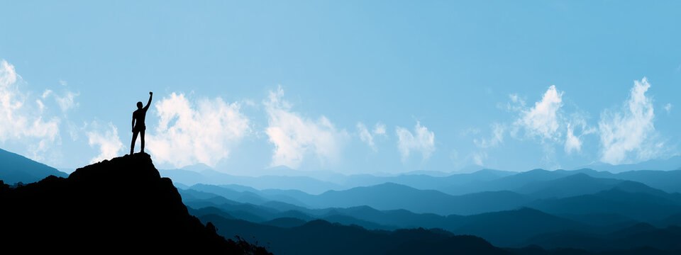 Silhouette of a triumphant man standing on a cliff edge with raised arms, overlooking a beautiful blue mountain range, symbolizing success, freedom, and adventure