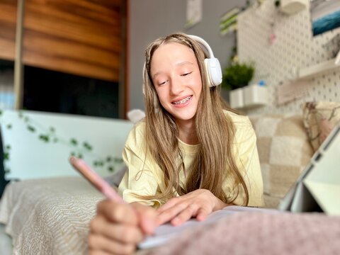 Teen girl with braces studying online at home, reading a book for school project. Modern distance learning and e‑education concept.