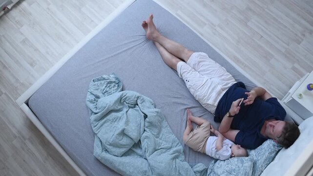 baby sleeps next to dad on bed. man lies on bed, holds phone in hands, looks at it. sleeping baby son lies next to him. Top view	