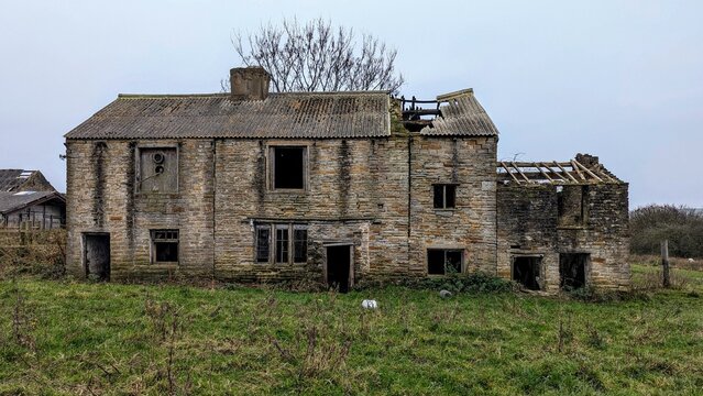 Abandoned collapsing country farmhouse, Lancashire UK. Derelict rural farm cottage building in grass field. Falling down countryside house. Uninhabited burnt out stone shell ruin, wrecked roof decay.